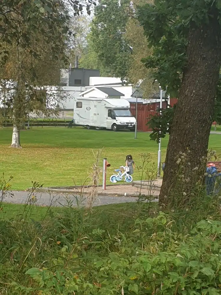 Bild av Sjöparkens camping visar en lummig parkmiljö med träd, gräsmattor och skuggiga områden. Fritidsfordon syns i bakgrunden, omgivna av ett välskött grönområde som inbjuder till rekreation och avkoppling.