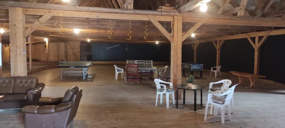 Bild av en rustik inomhusmiljö vid Abrahams Camp med trägolv och tak. Rummet innehåller trästolar och bord arrangerade som i en restaurang, med taklampor som ger en varm belysning.