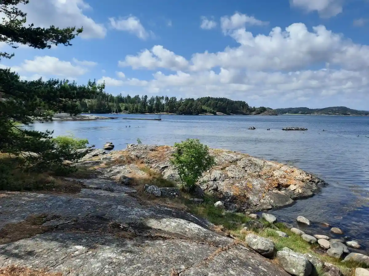 En klarblå himmel speglas i det lugna vattnet vid Almöns bad & camping, omgiven av klippor och kustlandskap. Några vita moln svävar över horisonten.