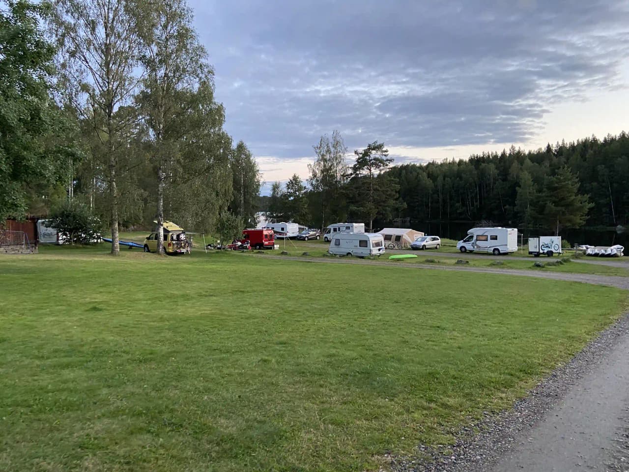 En grönskande campingplats vid Vammervikens camping med träd och gräs, flera husbilar parkerade, omgivna av ett öppet landskap under en molnig himmel.