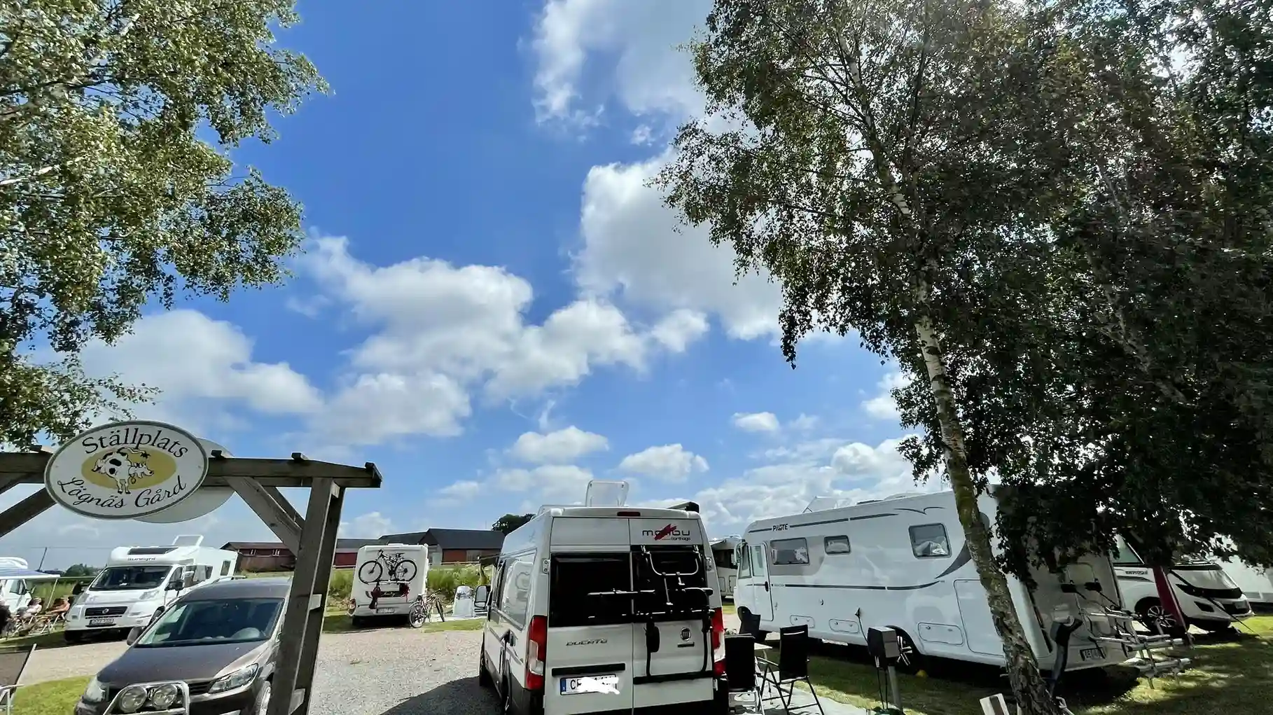 Bild av parkeringen vid Hotell & Spa Lögnäs Gård, med flera bilar och husbilar parkerade på området. Blå himmel och klart väder i bakgrunden.