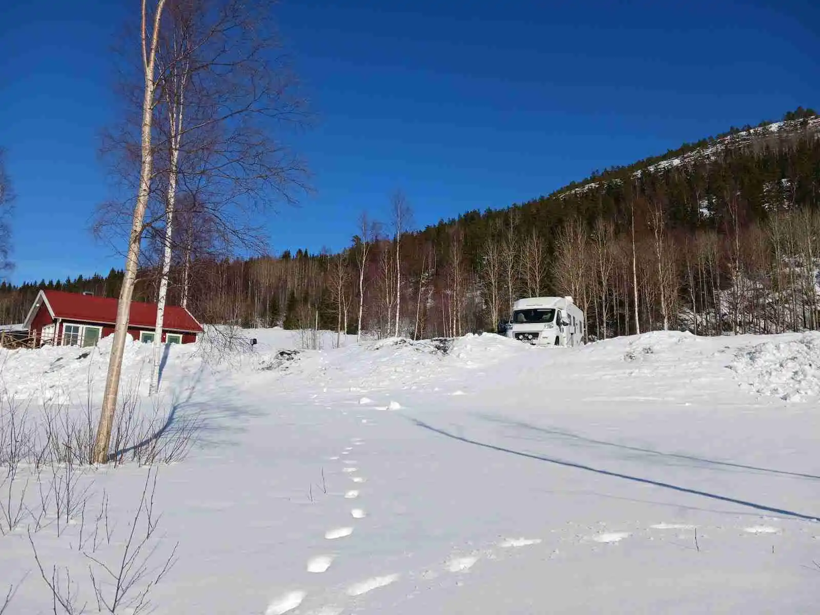 En snötäckt ställplats vid Gällstasjön omgiven av vinterlandskap med träd och lätt kuperad terräng.