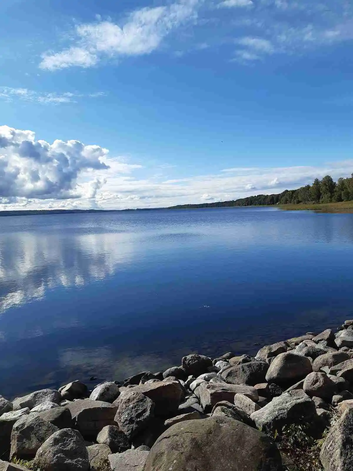 Utsikt över Vegby camping med blå himmel, moln och en vattenyta i förgrunden, omgiven av naturlandskap.