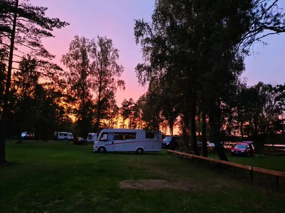 En husbil parkerad vid Orrefors camping i skymningen, omgiven av grönska och med svagt solsken i bakgrunden.