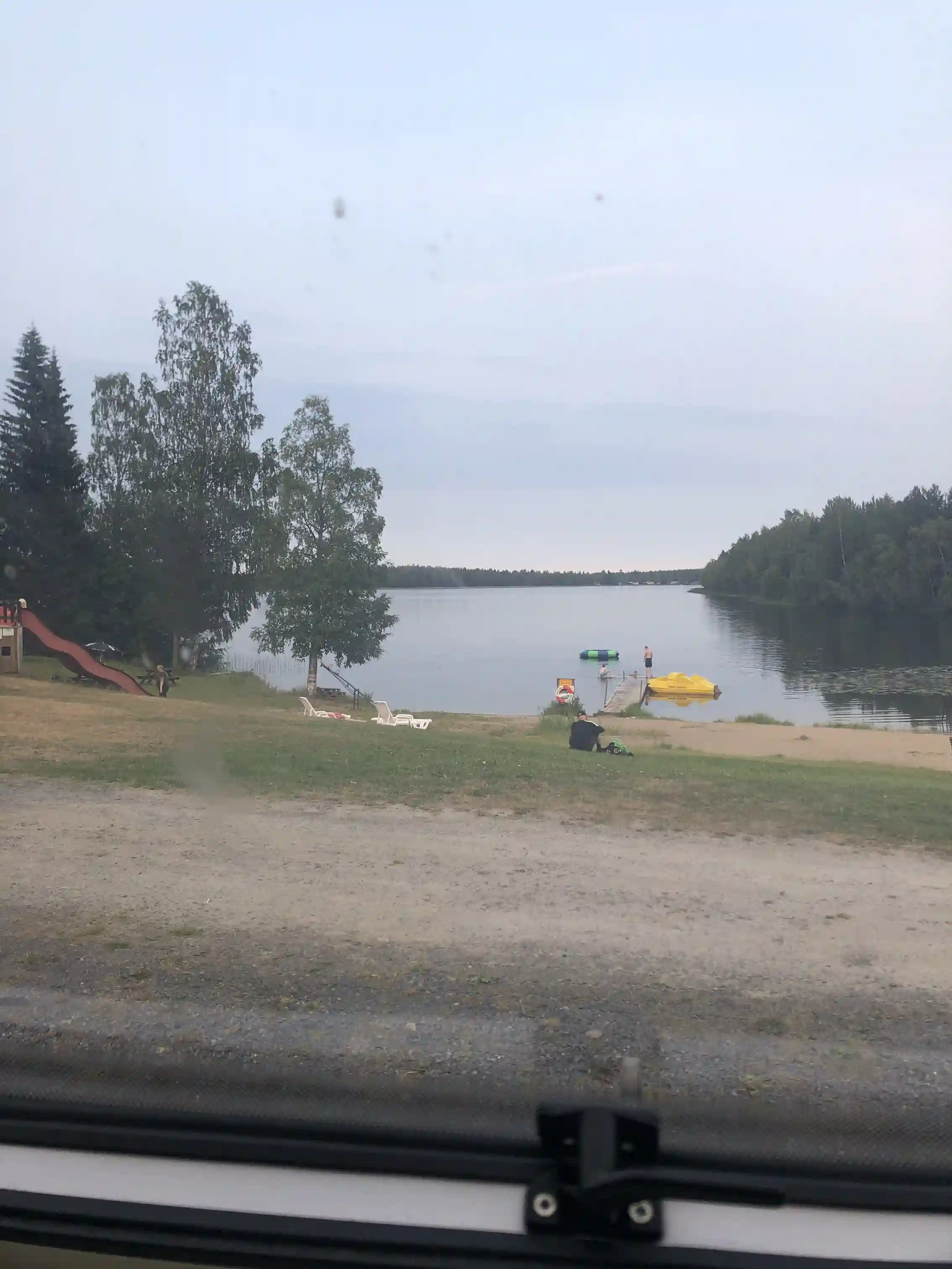Bild av Mosjöns camping: en naturskön sjö omgiven av grönskande skogar, under en delvis molnig himmel. I förgrunden syns en klassisk kit car parkerad vid sjön, vilket ger en nostalgisk och äventyrlig känsla till platsen.