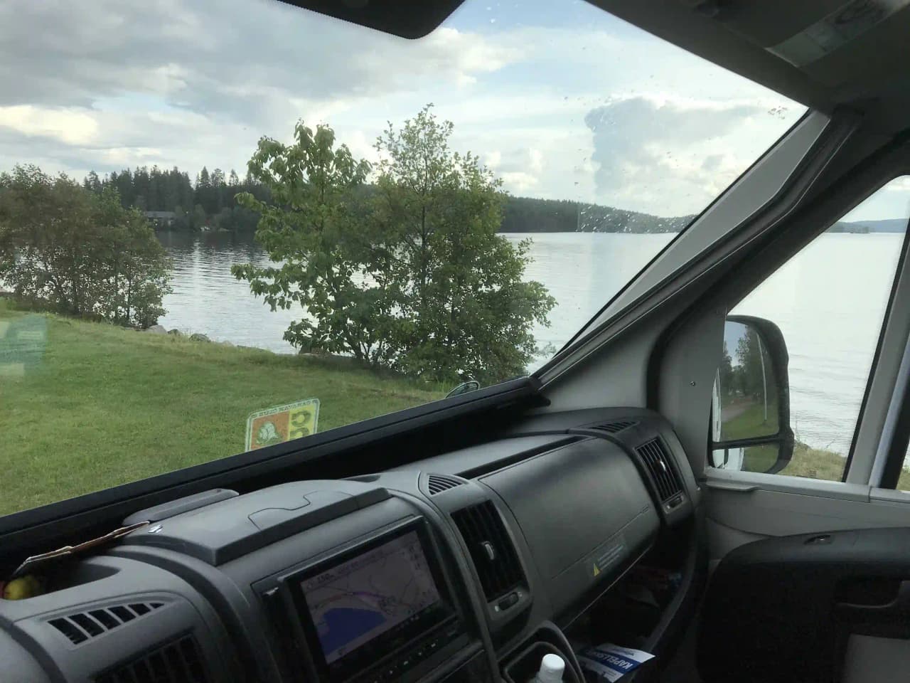 En husbil parkerad vid Ekeby Camping, med förarhyttens fönster och backspegel tydligt synliga. Bakgrunden visar campingen med grönska och en klar himmel, vilket ger en känsla av en trevlig plats för en roadtrip.