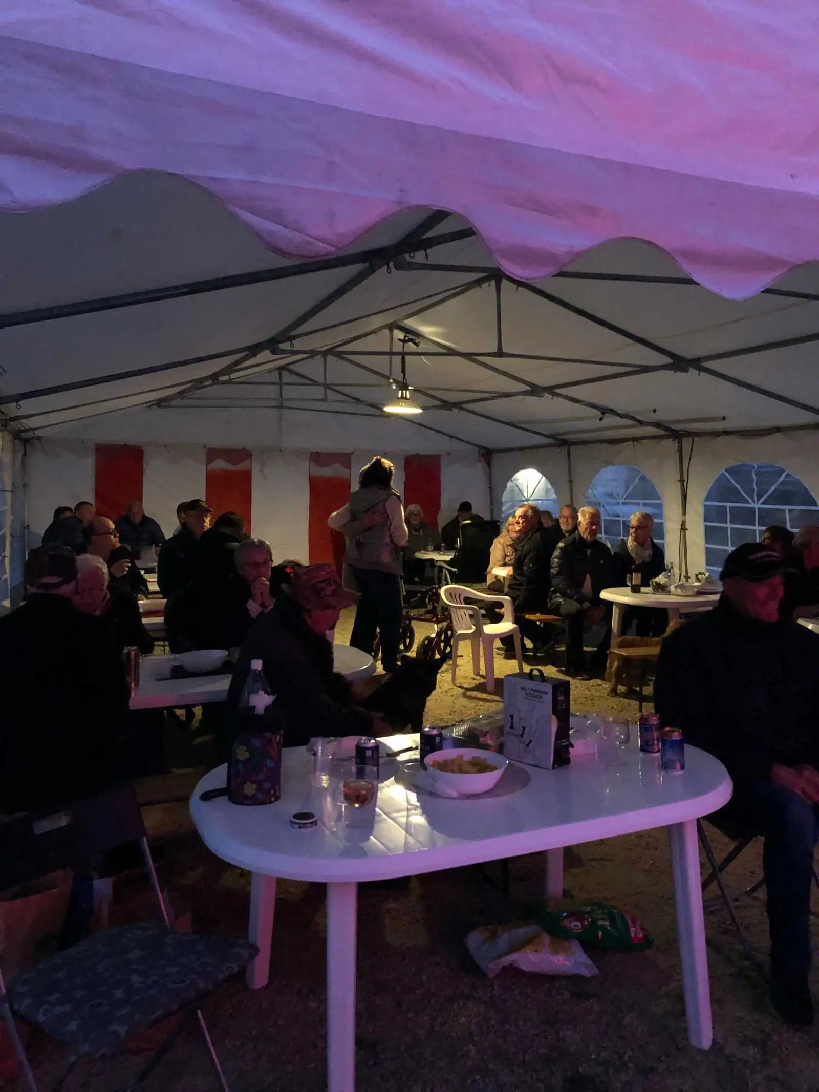 En bild av Kerstins camping som visar ett tält med tillhörande utemöbler, inklusive bord och stolar under ett soltak. Flera personer sitter i en avslappnad konversation, och atmosfären är avslappnad och trevlig med ett fokus på utomhusaktiviteter och gemenskap.