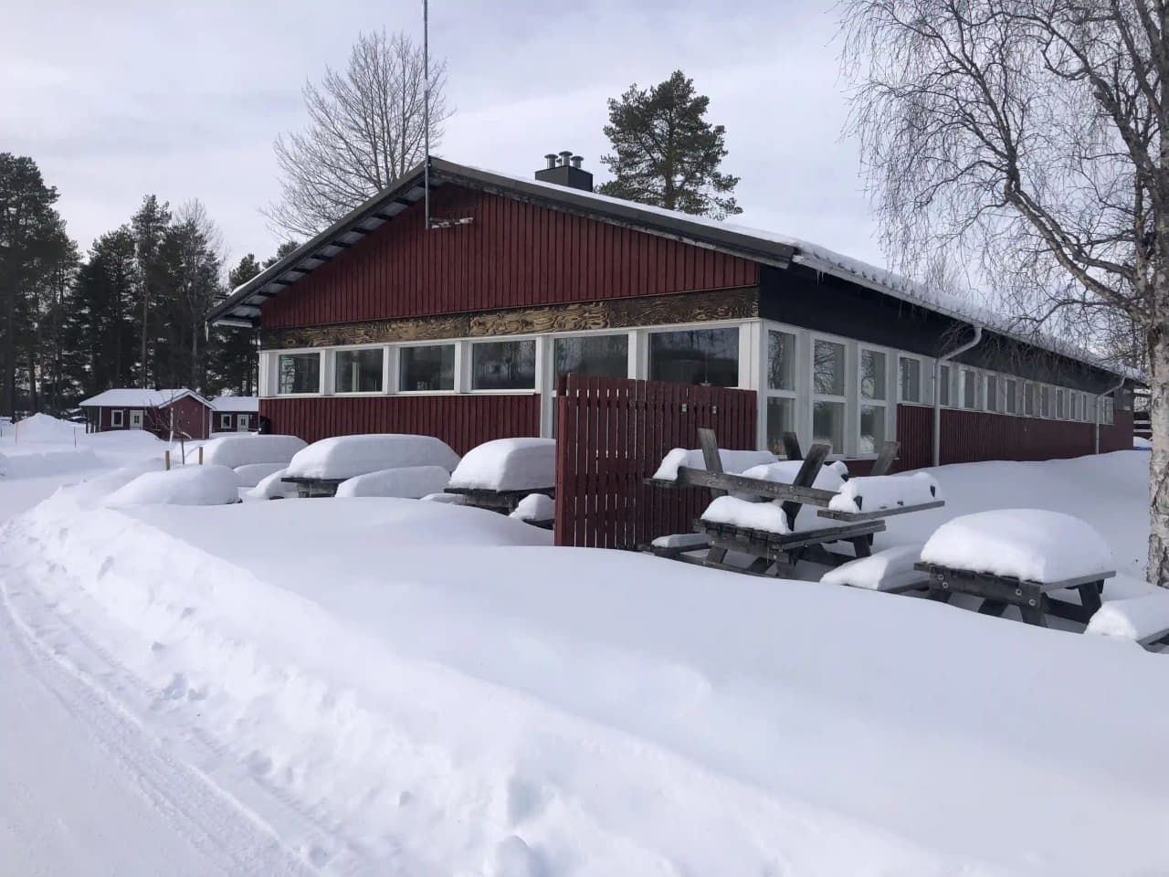 En snötäckt stuga vid Storuman Camping under vintern, med grenar böjda av snö och ett isigt tak.