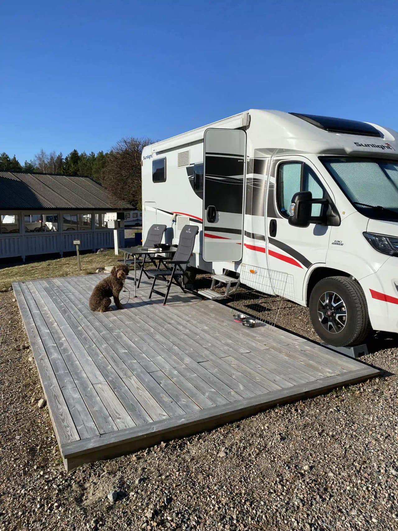 En campingplats med en husvagn och en husbil parkerade, en cykel står lutad mot en träplanka bredvid fordonen.