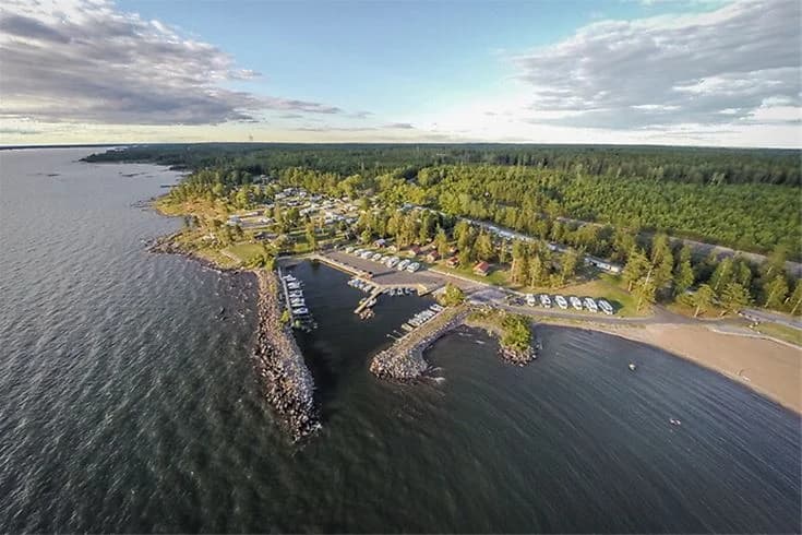 Bild av en kustlinje vid Askeviks Camping & Stugor, med en strand och en småbåtshamn omgiven av vatten i en vik eller kanal.