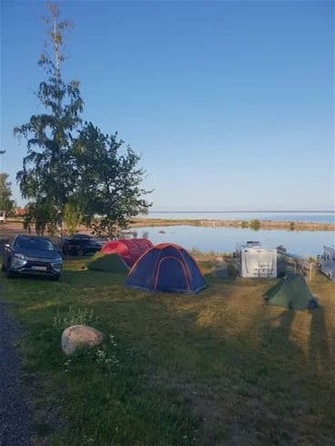 En campingplats nära kusten med tält och presenningar uppsatta. En sport utility vehicle är parkerad i bakgrunden, och campingutrustning syns runtom tältplatserna vid strandkanten.