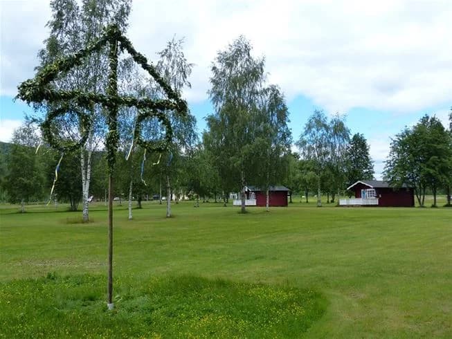 En grönskande campingplats med vidsträckt gräsmatta och omgivande träd i vårsol.