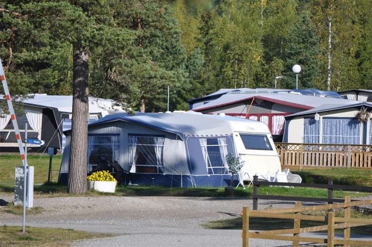 En campingplats med flera husbilar och husvagnar parkerade i skuggan, omgivna av gröna ytor. Några tält syns också, och campingområdet har en avslappnad atmosfär.