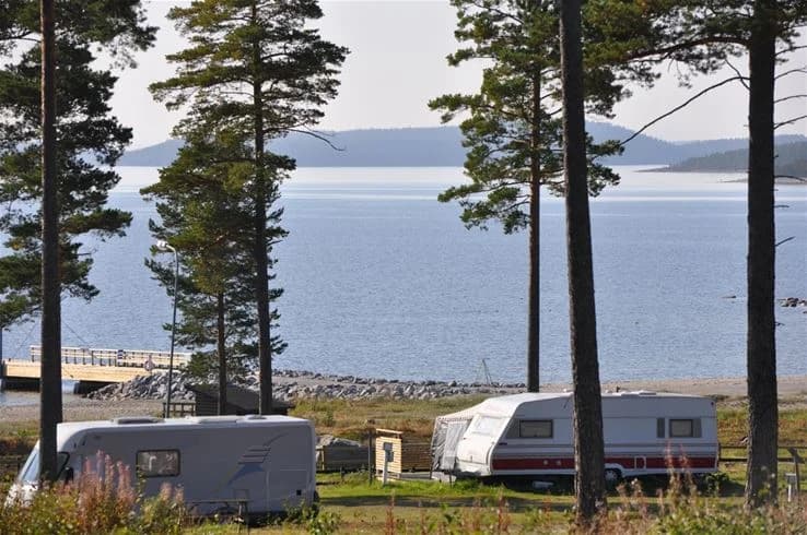 Vy över kustlandskapet vid Gullviks havsbad camping & stugby med en strandlinje i förgrunden, ett par husvagnar uppställda och en glittrande sjö eller havsvik i bakgrunden.