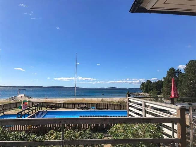 En solig dag vid Gullviks Havsbad Camping & Stugby med en pool omgiven av solstolar och parasoller. I bakgrunden syns en träpromenad och campingens stugor. Perfekt semesterort för avkoppling och nöje vid havet.