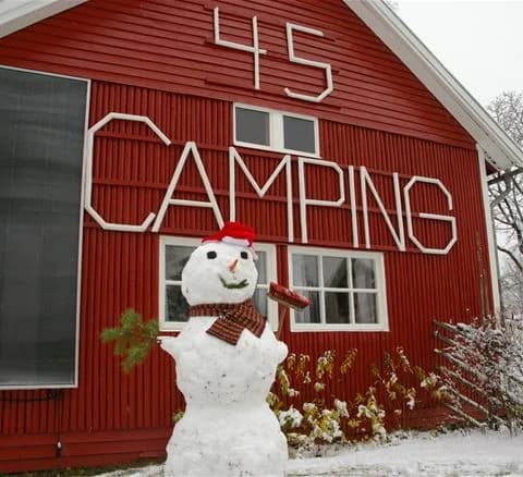 En snötäckt campingplats vid Camping 45 med en timmerstuga omgiven av vinterlandskap. Marken är täckt av snö och stugans sidoväggar är delvis täckta av vitt, vilket skapar en mysig atmosfär.