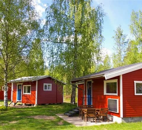 Alt-text: En campingplats i landsbygden med en stuga och en log cabin omgivna av grönskande gräsmattor och trädgårdsbyggnader.