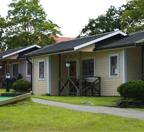 Bilden visar Villa Björkhagen, en charmig stuga med grönmålad fasad och vita knutar. Omgiven av en välskött trädgård med grönskande gräsmatta och en stensatt uppfart.