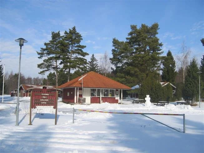 En snötäckt campingplats vid Kapellskär med små stugor som omges av träd. Taken är täckta av snö och en flaggstång står bredvid en av stugorna. Vinterlandskapet ger en fridfull och avkopplande atmosfär.