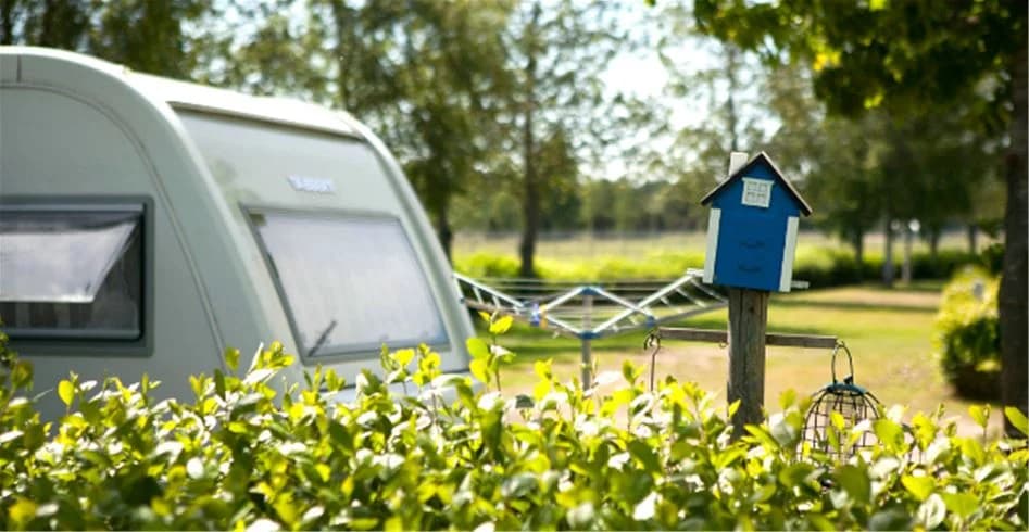 En husvagn uppställd på en campingplats omgiven av grön natur, med soligt väder i bakgrunden.