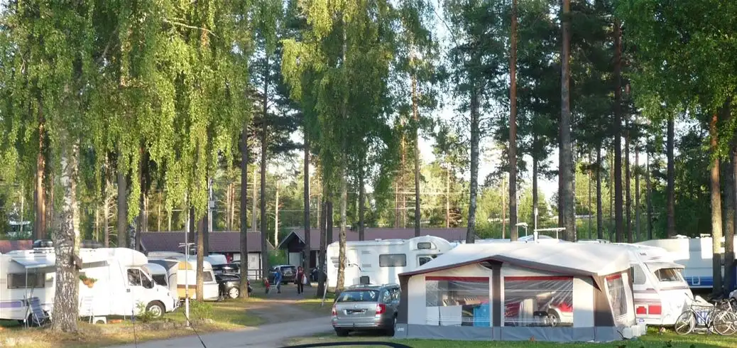 En husvagn är parkerad vid en campingplats omgiven av gröna barrträd. I förgrunden syns en öppen yta med parkeringsmöjlighet.