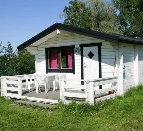 En vinterlandskap med flera timmerstugor på Gustavsvik Resort, omgivna av snö och träd. Varje stuga har ett perfekt placerat sadeltak och träfasad, vilket smälter in harmoniskt med den naturnära omgivningen.