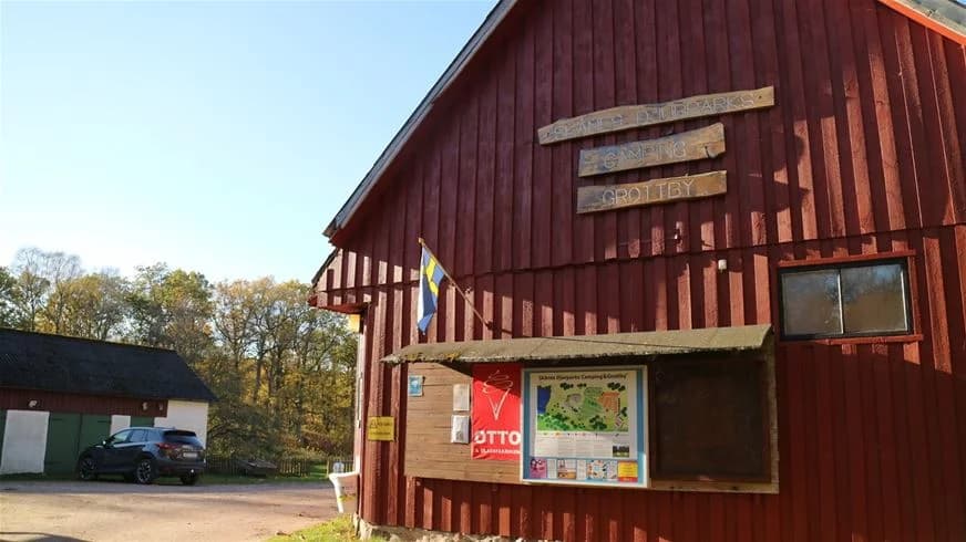 En träbyggnad med röd fasad och vita knutar, vilket ger intryck av en traditionell svensk lada. Marken omkring byggnaden är omgiven av grönska och ett naturskönt landskap som speglar grottbyns camping.
