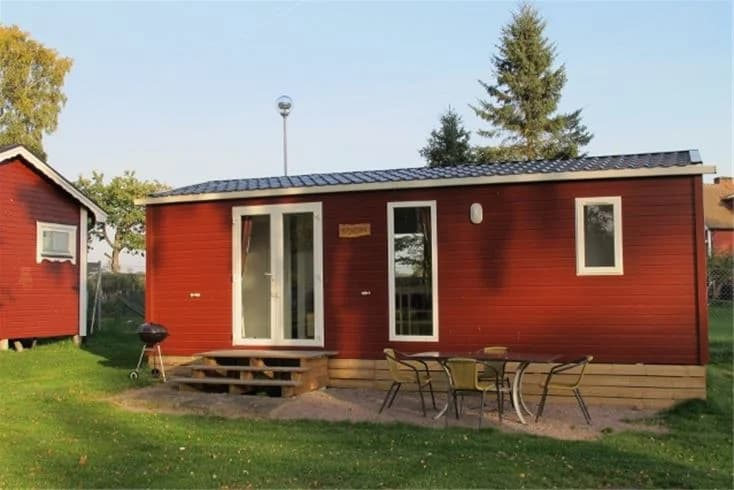 En campingstuga med träpanel omgiven av en grön gräsmatta på Jägersbo Camping. I bakgrunden syns trädgårdsbyggnader och en vedbod.