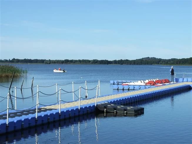 En vy över en blå sjö med brygga och båtar vid Jägersbo Camping Swecamp, omgiven av grönska.