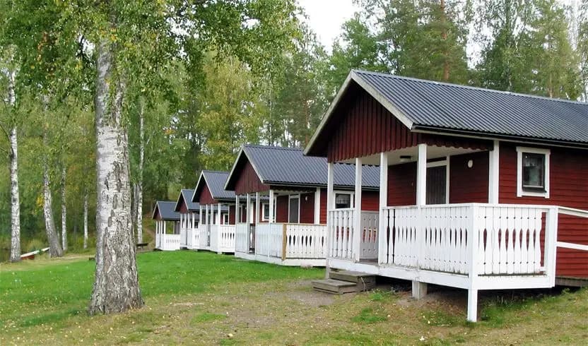 En bild av en timmerstuga med rött tak och en veranda, omgiven av grönskande gräsmatta på Degernäs Camping.