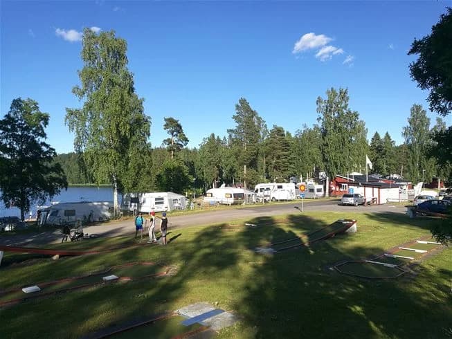 En grönskande campingplats med träd och växter omger öppna ytor där husvagnar är uppställda. Gröna gräsmattor sträcker sig vid sidan av platser för rekreation i det allmänna området på Degernäs camping.