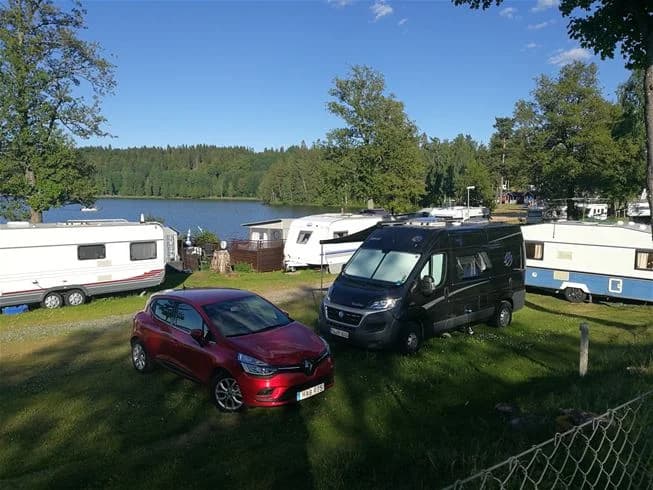 En campingplats med parkerade husbilar, husvagnar och personbilar på en öppen grusparkering vid Degernäs Camping. I bakgrunden syns skog och en klar himmel.
