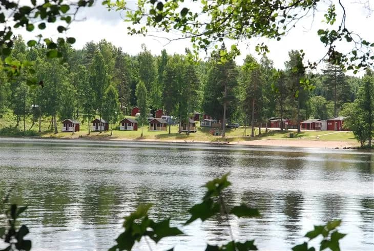 En pittoresk vy över Degernäs camping med en sjö i förgrunden, omgiven av träd och grönska. I bakgrunden syns en stuga vid vattenkanten.