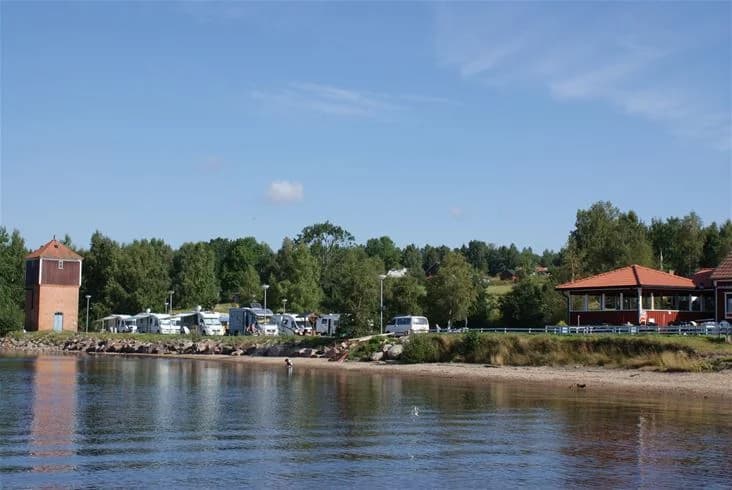 En lugn sjö omgiven av grönska vid Sandaholm Restaurang & Camping, med strand och trädkantad kustlinje i bakgrunden.