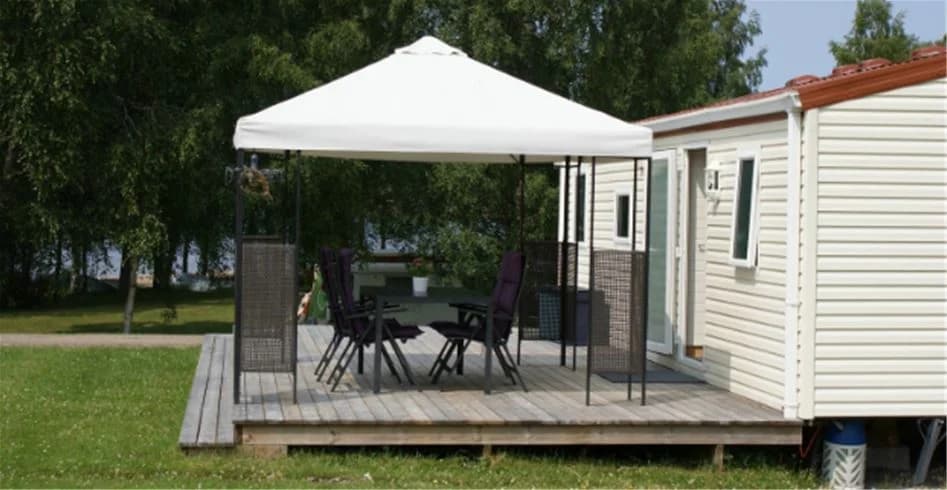 Skuggig uteplats på Sandaholm restaurang & camping med trädeck och markis, omgivet av grönska och mobilt hem i bakgrunden.