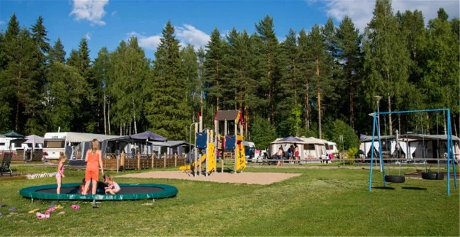 En grönskande campingmiljö med tält bland träd i en park. Människor njuter av fritidssysslor och avkoppling i skuggan, med stora gräsmattor och offentliga ytor runtomkring.
