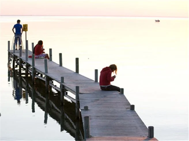 En brygga som sträcker sig ut i havet vid Lundegård Camping & Stugby. I förgrunden syns människor som promenerar och njuter av naturen, vilket förstärker känslan av semester och avkoppling.