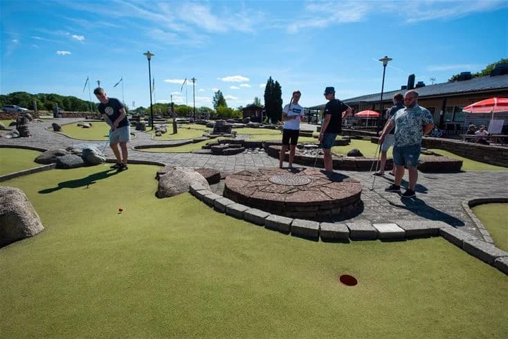 En solig dag på Lundegård Camping & Stugby med gäster som spelar minigolf i en välskött park. Området är fyllt av glädje och gemenskap, med besökare i olika åldrar som njuter av fritidsaktiviteter.
