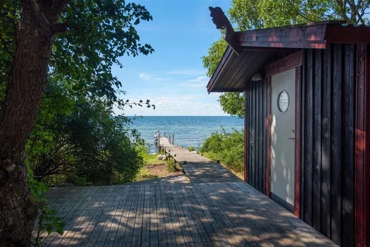 En träpromenad längs kusten vid Lundegård Camping & Stugby, omgiven av havet och skuggiga träd.