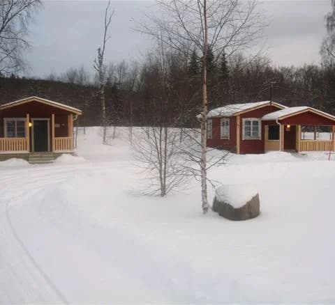 En vintrig scen vid Klarälvens camping med snötäckta träd och mark. En liten stuga med sluttande tak ses mitt i snön, vilket ger en känsla av stillhet och vinteridyll.