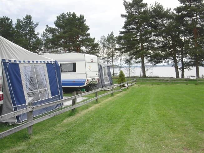 En husvagn parkerad på en grön gräsmatta i skuggan av träd på Malexander camping, med flera andra mobila hem och husbilar i bakgrunden.