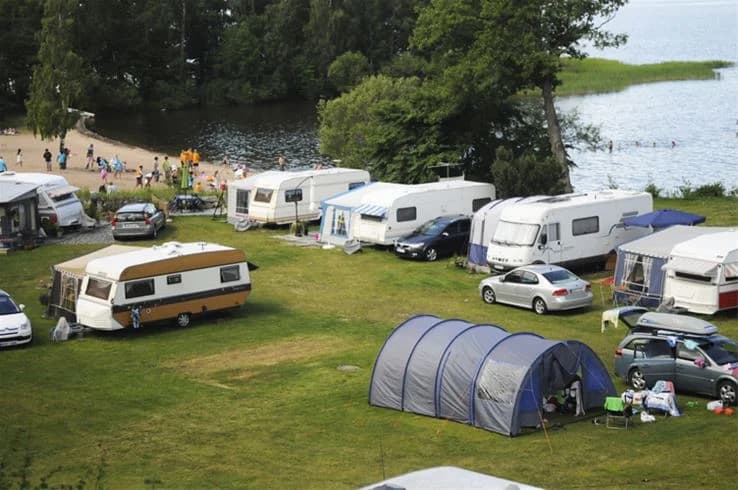 Bild av en campingplats vid stranden med tält uppställda, omgiven av grönska och en sjö i bakgrunden, som visar en avkopplande och naturskön miljö på Hjälmargården & Läppe Camping.