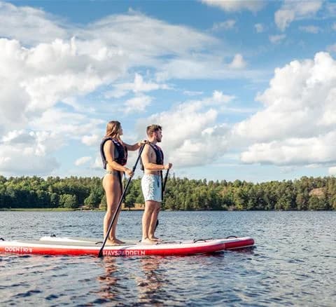 Personer som paddelsurfar och njuter av vattensporter vid Gålö Havsbad. Lekfull och avkopplande atmosfär vid stranden.