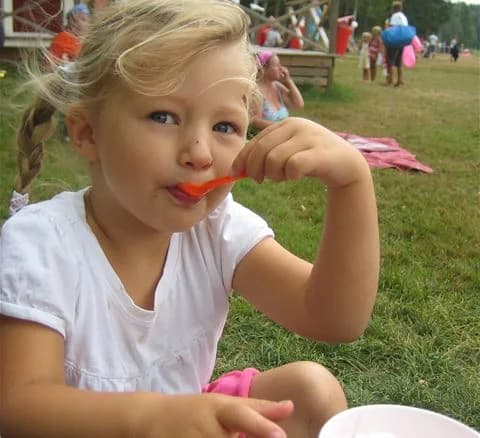 Ett ungt barn med blont hår njuter av en glass på en solig sommardag vid Gålö Havsbad. Barnet ser glatt ut medan det håller glassen i handen och tar en tugga. I bakgrunden syns strandmiljön som ger en inbjudande sommarkänsla.