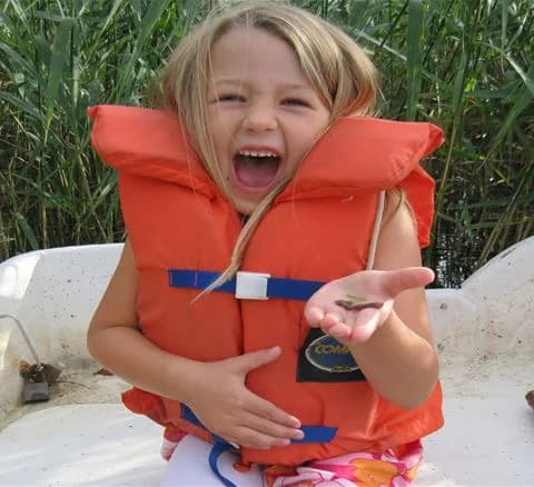 Ett småbarn iklädd en orange flytväst leker vid kanten av vattnet på Gålö Havsbad. Barnet ser glatt och förväntansfullt ut bland sand och vatten, vilket fångar en känsla av somrig semester och lekfullhet.