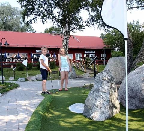 Ett grönt område vid Gålö havsbad med människor som njuter av fritidsaktiviteter, som minigolf och parklek i det soliga vädret.