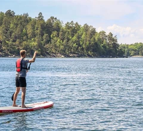 En person som glider fram på en standup paddleboard på det klara vattnet vid Gålö havsbad, omgiven av vacker natur och sol. Perfekt för friluftsaktiviteter och att uppskatta skärgårdens skönhet.