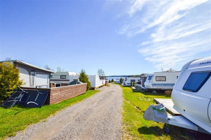 En campingplats vid kusten på Charlottenberg camping & stugor - Haganäset. En husvagn står parkerad nära stranden med utsikt över en vik, omgiven av grönt landskap och vatten.
