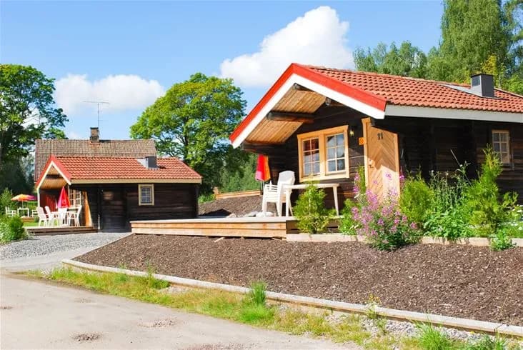En mysig timmerstuga omgiven av grönska, belägen på Charlottenberg camping & stugor - Haganäset. I bakgrunden syns fler stugor och träd, vilket skapar en inbjudande och lantlig atmosfär.