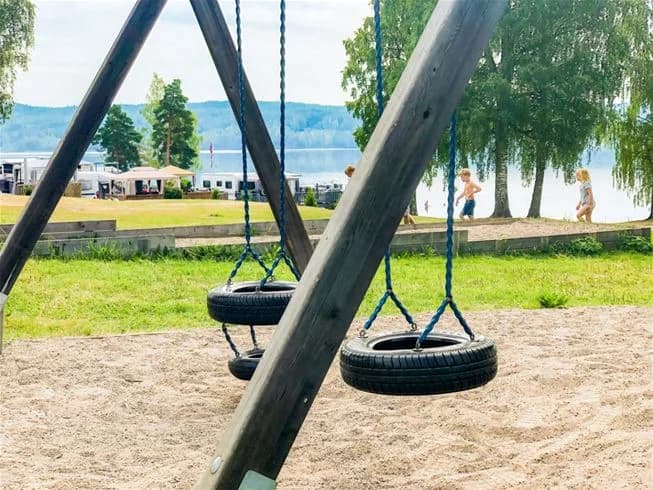 En lekplats på Charlottenberg camping & stugor - Haganäset, med gungor, klätterutrustning, och skuggiga sittplatser, där barn och familjer njuter av utomhuslek och rekreation.